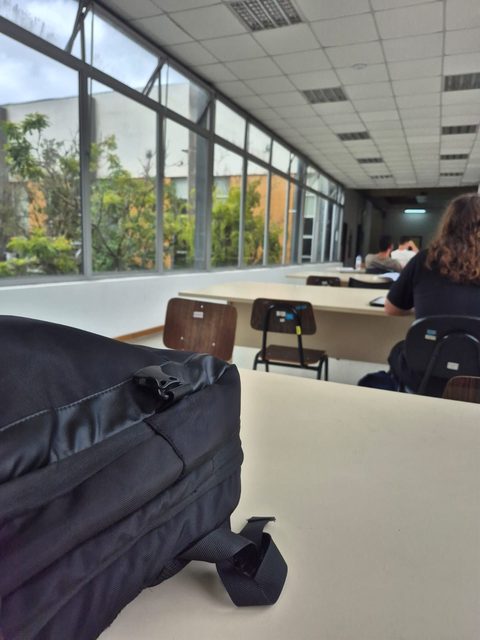 Mochila sobre uma mesa em primeiro plano, com uma sala de aula desfocada ao fundo, mesas vazias e um corredor envidraçado.
